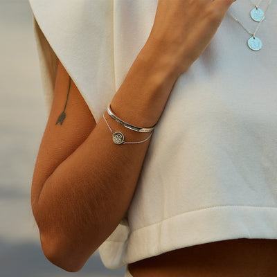 Inzoomad arm med två armband och en tatuering, kvinnan har även två halsband utanpå en vit tröja